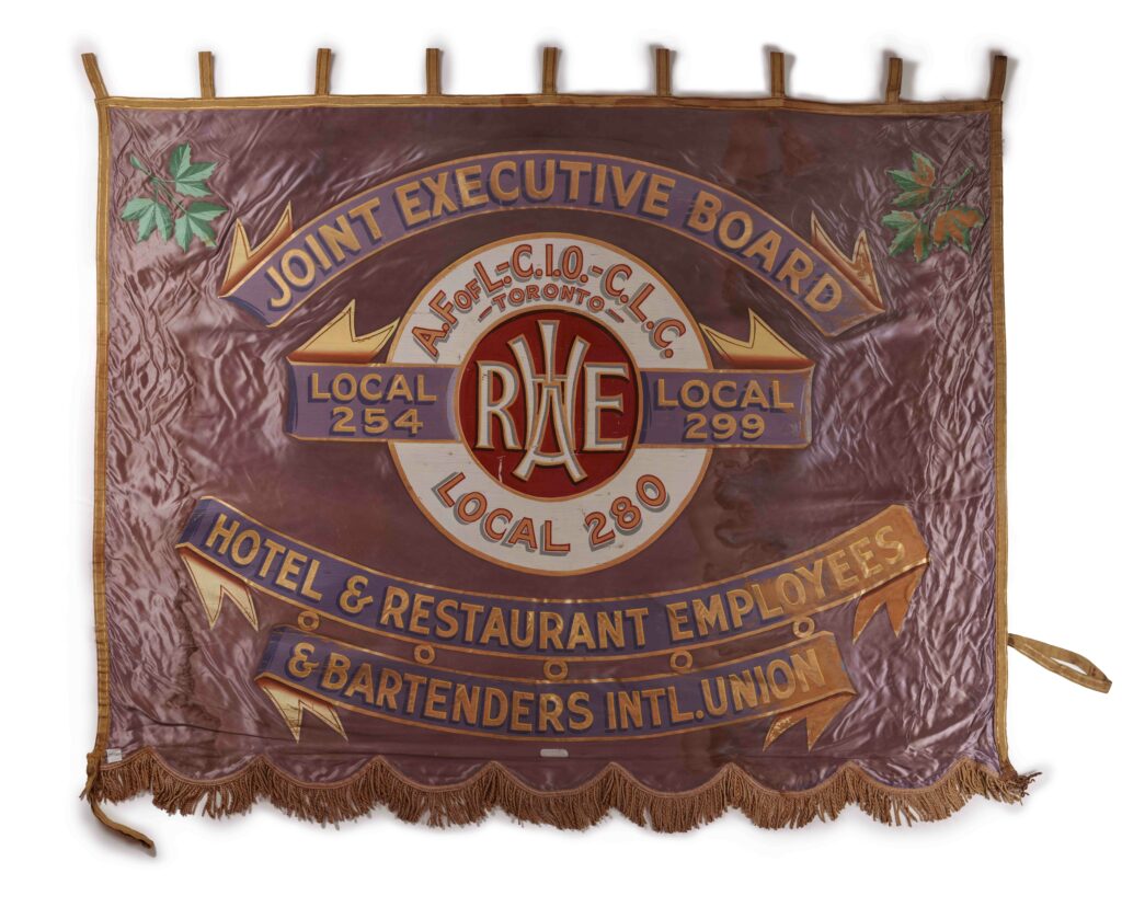 A shiny decorative banner with a purple background, a gold border, and blue decorative scrolls of text, for board, union and locals of the HOTEL & RESTAURANT EMPLOYEES & BARTENDERS INTL. UNION, and a red white and gold circular logo in the middle, gold tassels along the lower edge, gold tabs along the top.