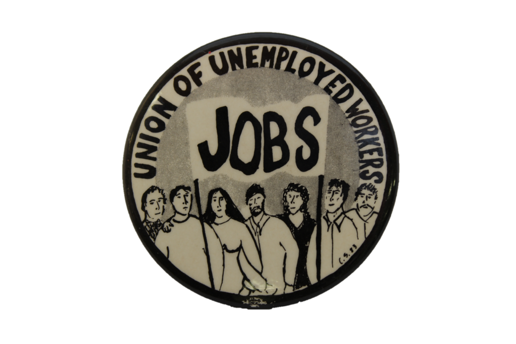 Black and white circular object with black edge, illustration of people carrying a banner with JOBS written on it, UNION OF UNEMPLOYED WORKERS written around the edge of the circle 