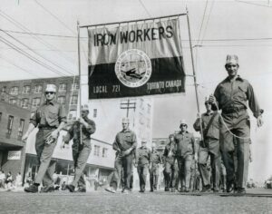 Black and white photo of men in uniform marching in the street, the two in front carrying a banner reading IRON WORKERS at the top