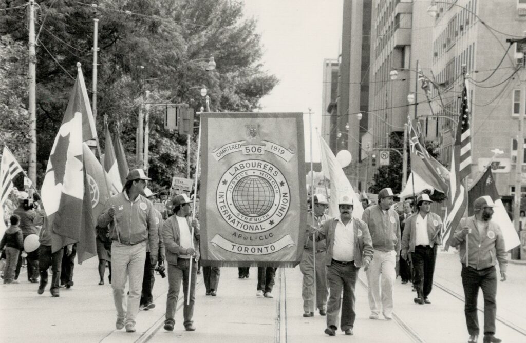 Black and white photo of parade of men in caps, two in front carry a union banner between them, behind the men carry many flags, including the American and Canadian national flags