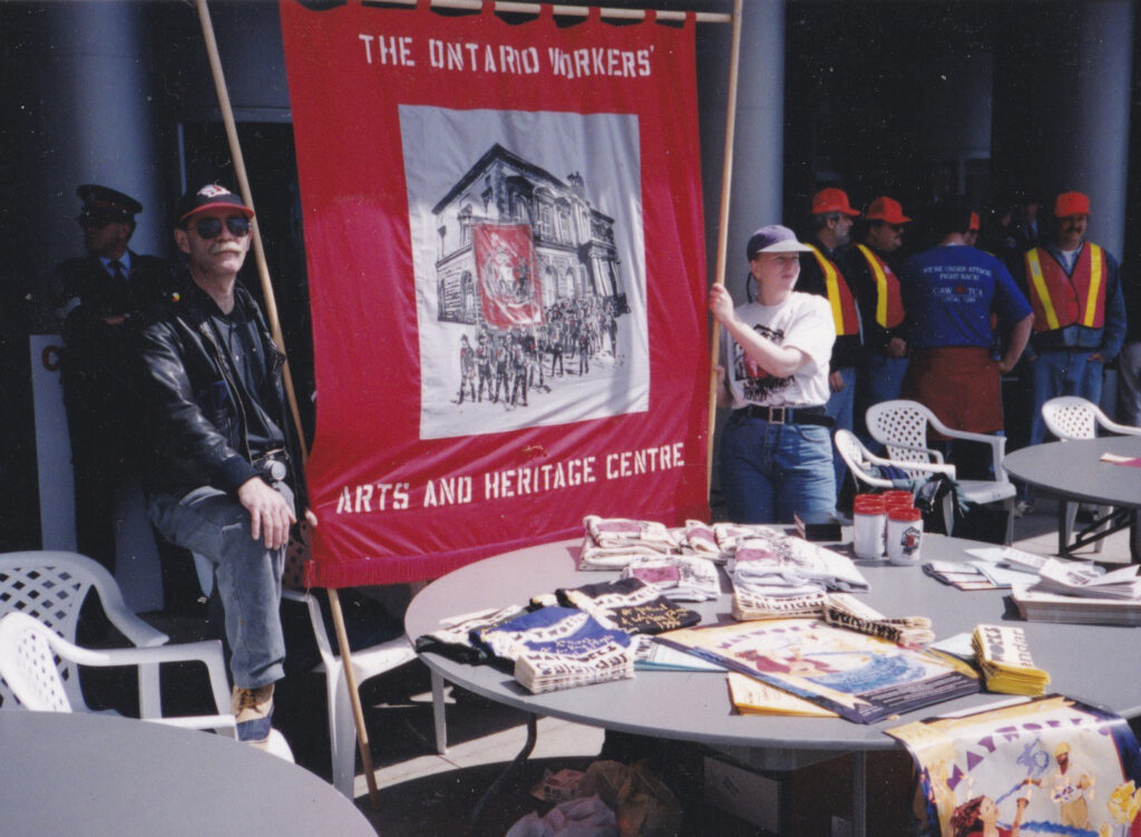 Colour photo of a man and a woman wearing baseball caps standing either side of a red banner they are holding, in the foreground is a table, behind the woman are a group of people in high visibility jackets and hard hats