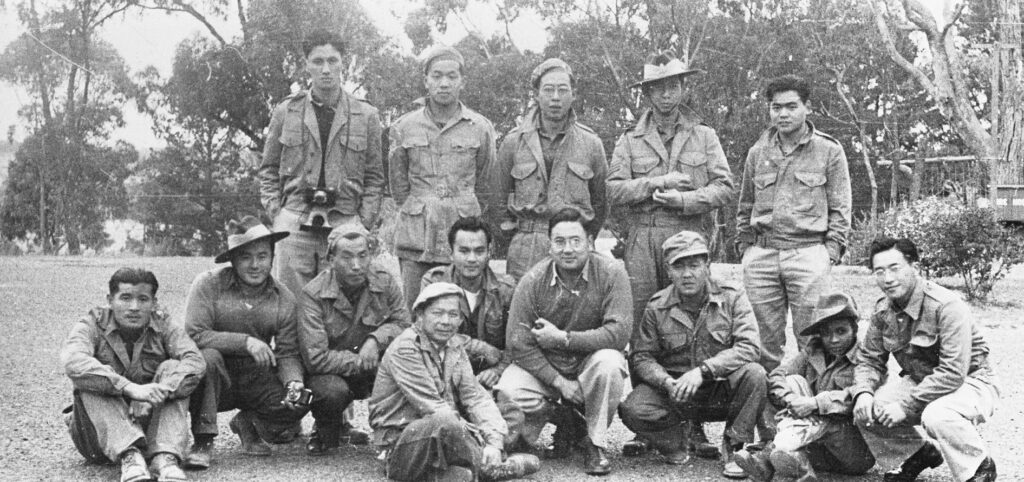 Black and white photo of 14 men, 5 standing behing the rest, who are seated on the ground, some wear hats or caps, one has binoculars around his neck